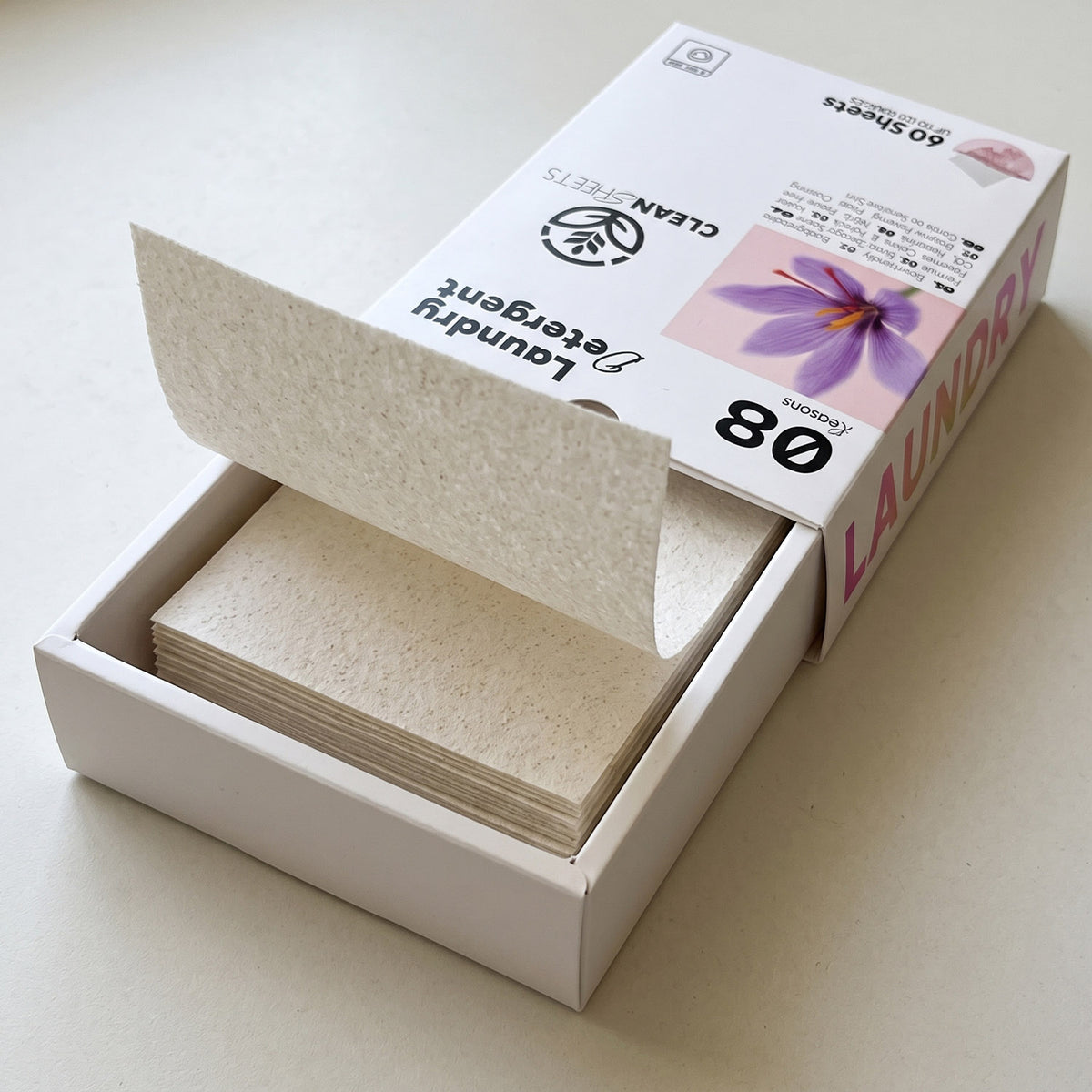 A box of laundry detergent sheets with the lid open, showing a stack of textured off-white sheets inside. The packaging is white with pink and black text and a purple flower graphic.